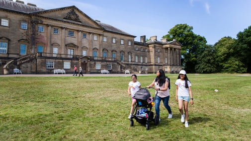 A group of people with a pushchair walk across a lawn in front of a mansion house